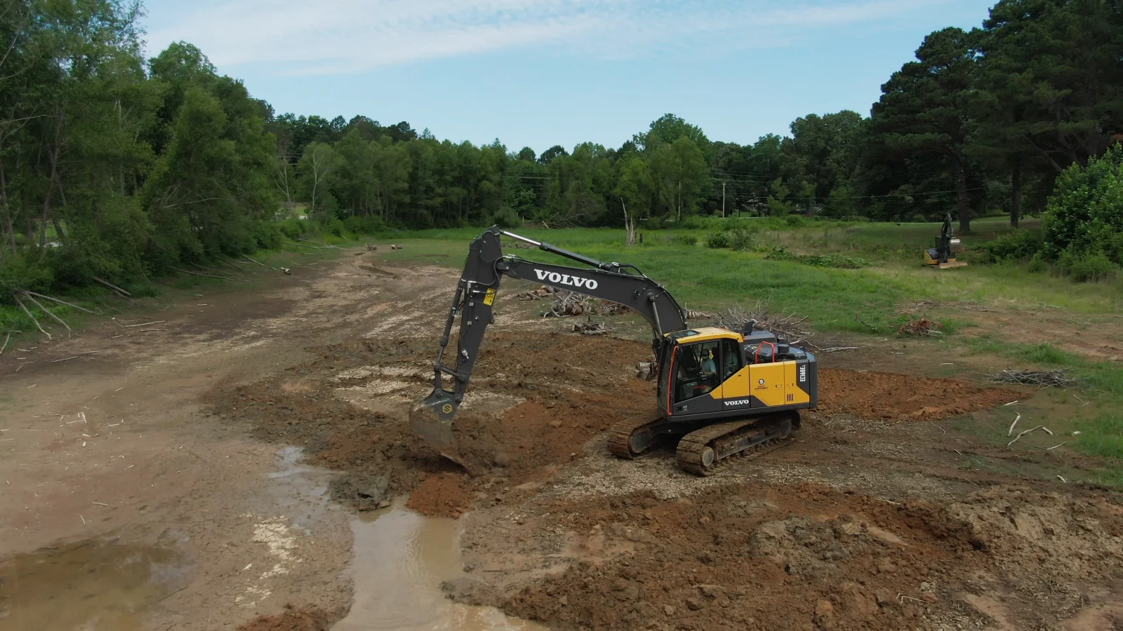 Excavator shaping a pond basin in an open field, with water pooling in the excavation.